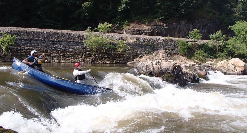 Two people wearing safety gear paddle a canoe over a small waterfall. 